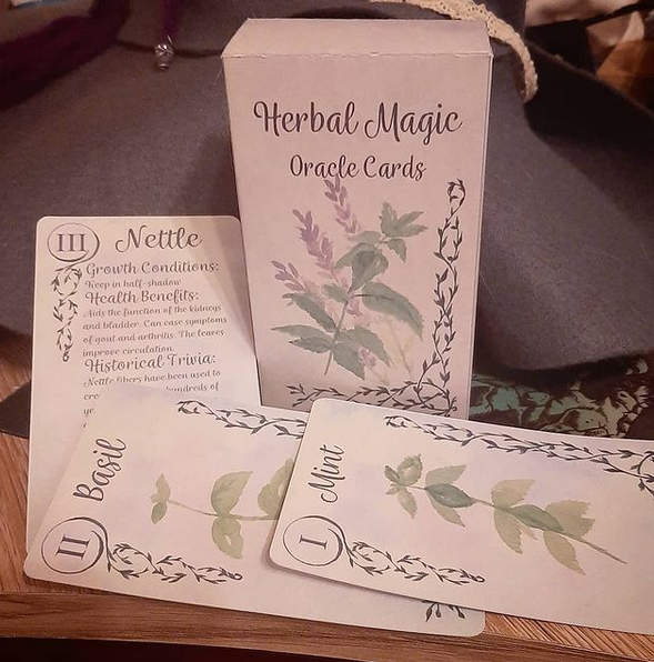 A picture of a box in Tarot format (12x7cm) with "Herbal Magic Oracle Cards" written on it. Underneath lie 3 cards. 2 have water colour paintings of herbs on them and say " 1 Mint" and "2 Basil" while the 3rd one is a backside saying "3 Nettle" as well as containing information on growth conditions, health benefits and historical trivia. The rest of the 3rd card is obscured by the others lying on top of it.